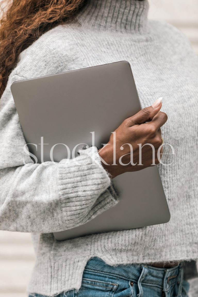 Vertical stock photo of a woman wearing a gray sweater holding a laptop computer.