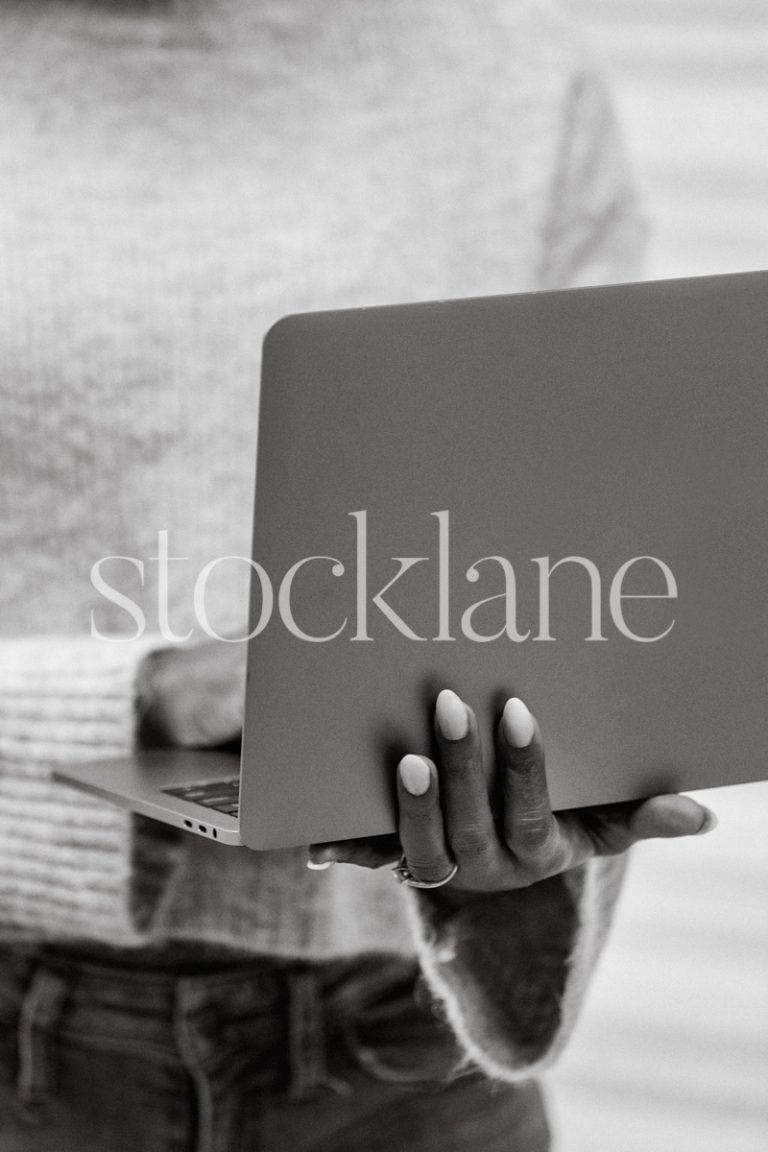 Vertical black and white stock photo of a woman wearing a gray sweater holding a laptop computer.