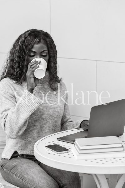 Vertical black and white stock photo of a woman having a cup of coffee while working on her laptop computer.