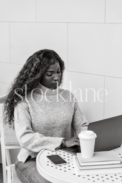 Vertical black and white stock photo of a woman having a cup of coffee while working on her laptop computer.