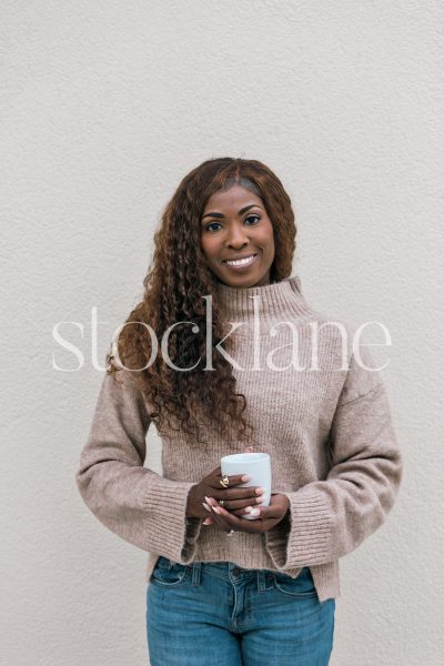 Vertical stock photo of a woman wearing a neutral colored sweater and jeans, holding a cup of coffee.