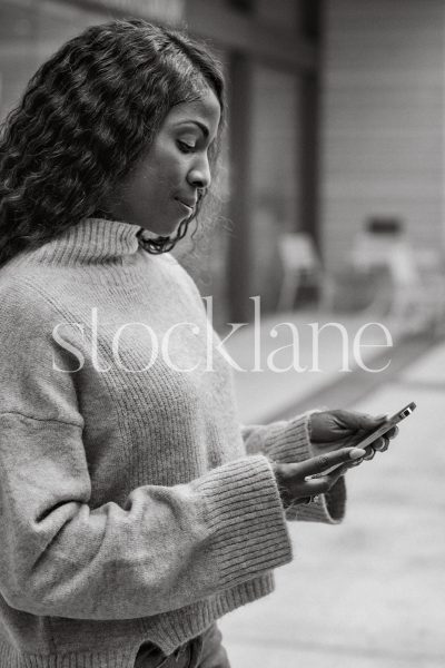 Vertical black and white stock photo of a woman wearing a neutral colored sweater, holding a phone.