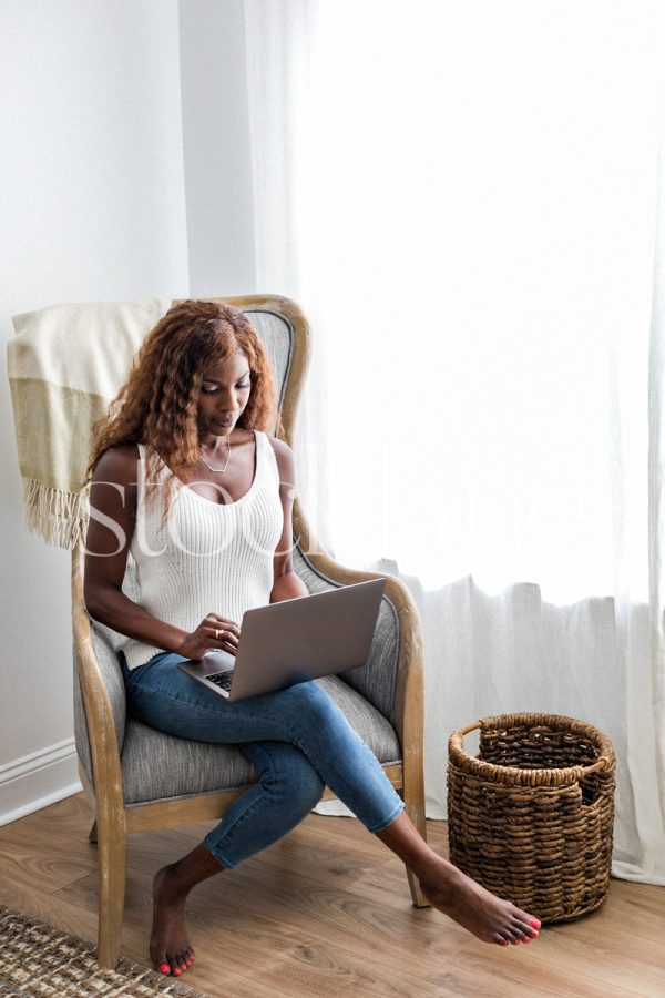 Vertical stock photo of a woman wearing a white top and jeans, sitting on an armchair working on her laptop computer.