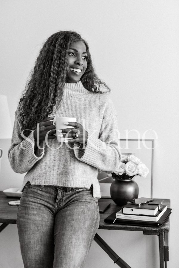 Vertical black and white photo of a woman holding a cup, leaning against her desk.