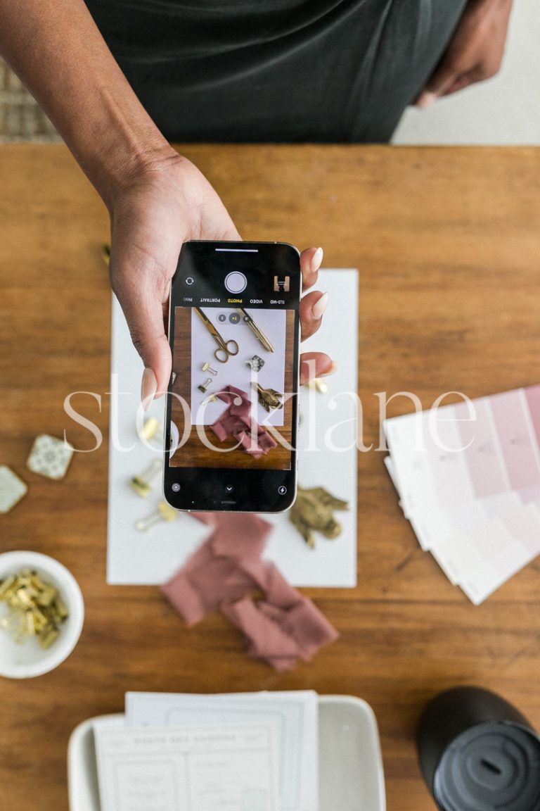 Vertical stock photo of a woman taking a photo of a flatlay on her desk with her phone.