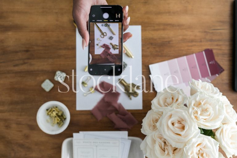 Horizontal stock photo of a woman taking a photo of a flatlay on her desk with her phone.