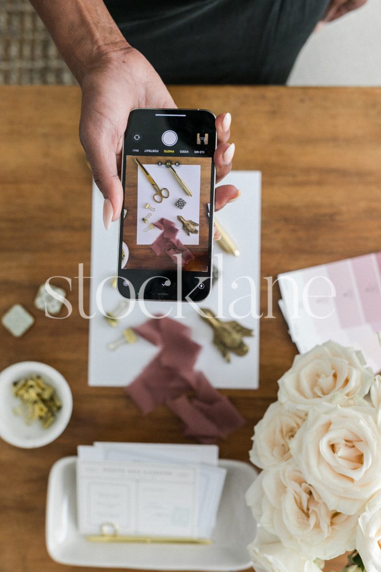 Vertical stock photo of a woman taking a photo of a flatlay on her desk with her phone.