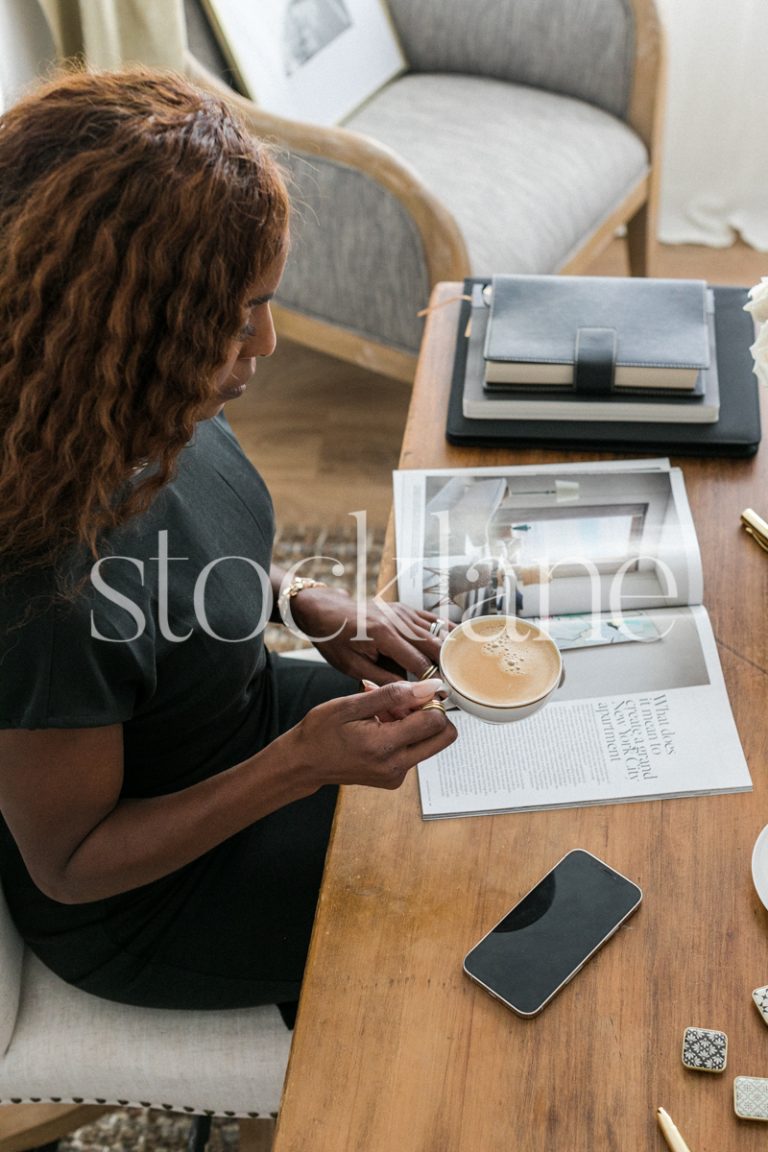 Vertical stock photo of a woman sitting at her desk, having a coffee and reading a magazine.