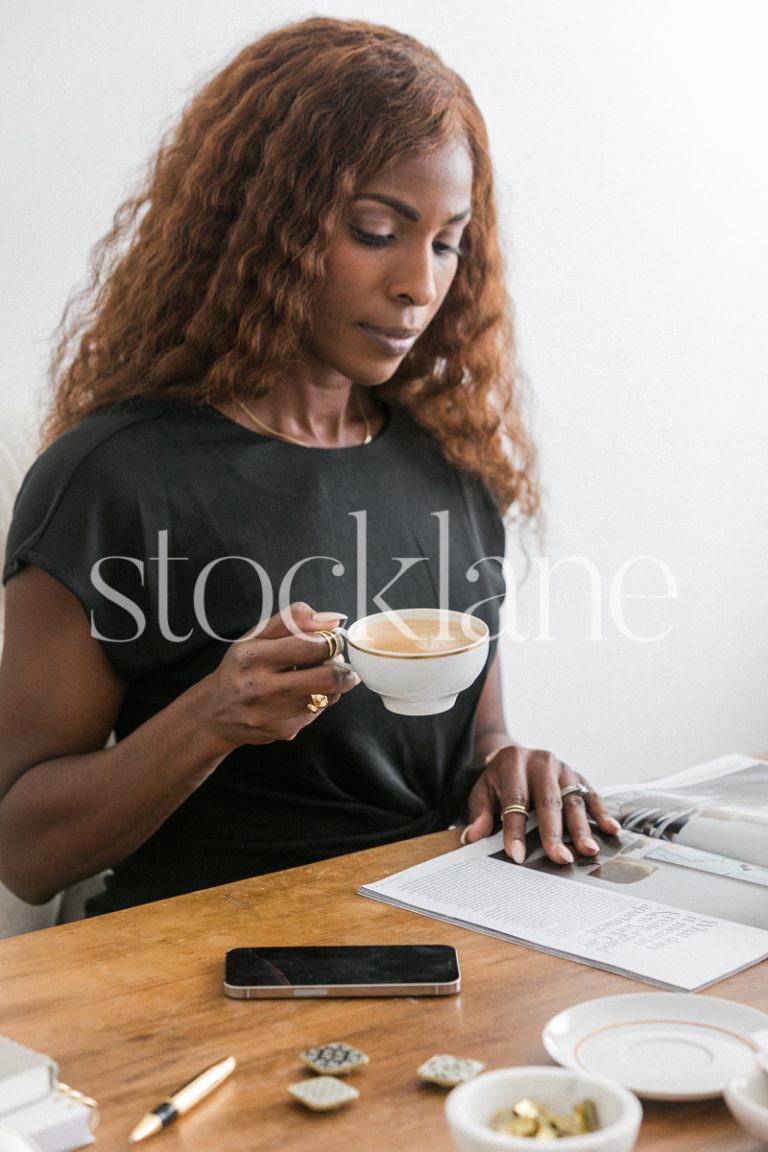 Vertical stock photo of a woman sitting at her desk, having a coffee and reading a magazine.