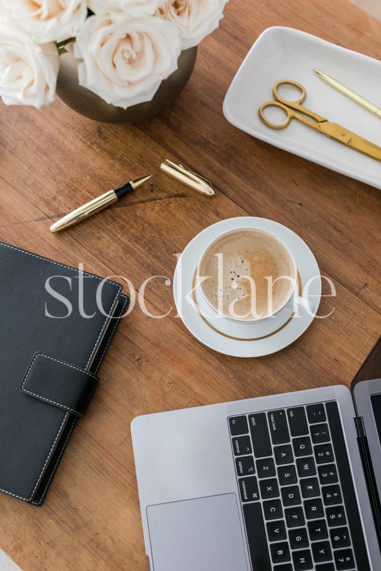 Vertical stock photo of a desk with a computer, a cup of coffee and other desk supplies, in gold and neutral colors.