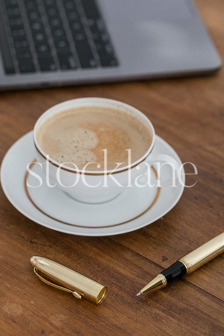 Vertical stock photo of a desk with a coffee cup, a computer and a gold pen.