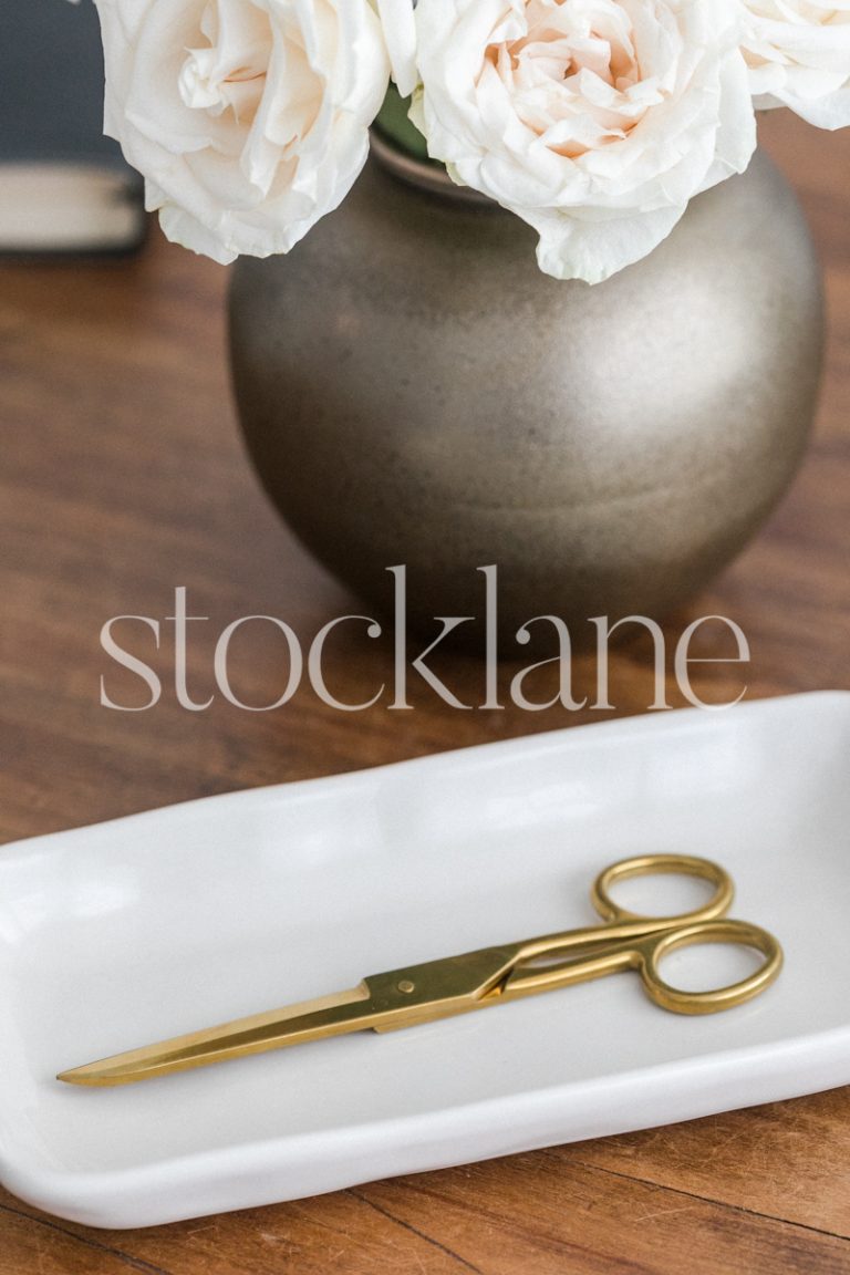 Vertical stock photo of gold scissors on a white tray, with flowers on the background.