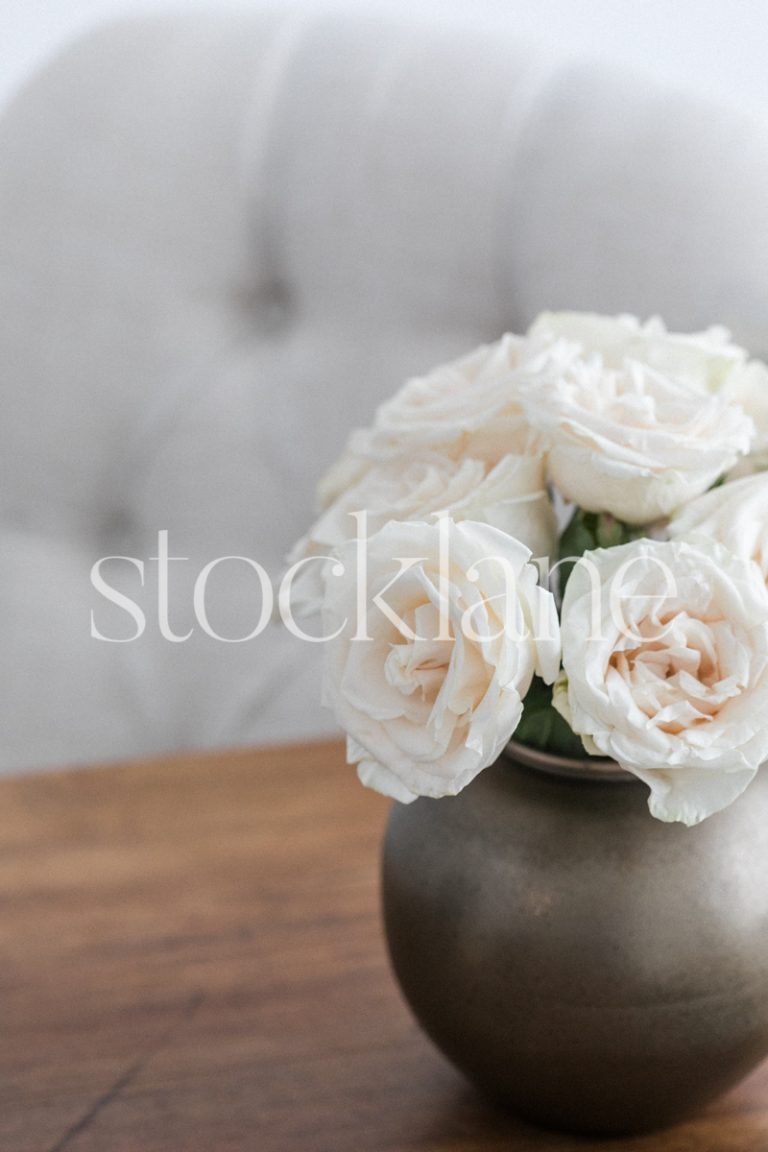 Vertical stock photo of a gold vase with white roses on a wood table.