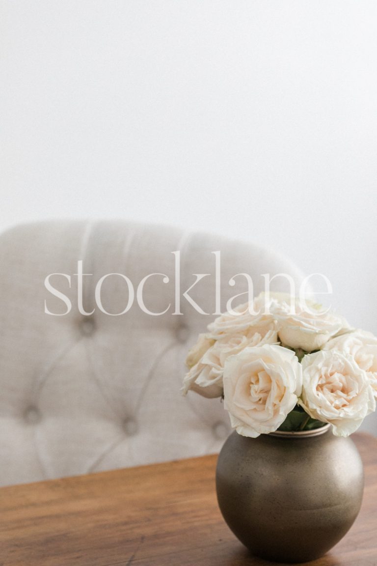 Vertical stock photo of a gold vase with white roses on a wood table.