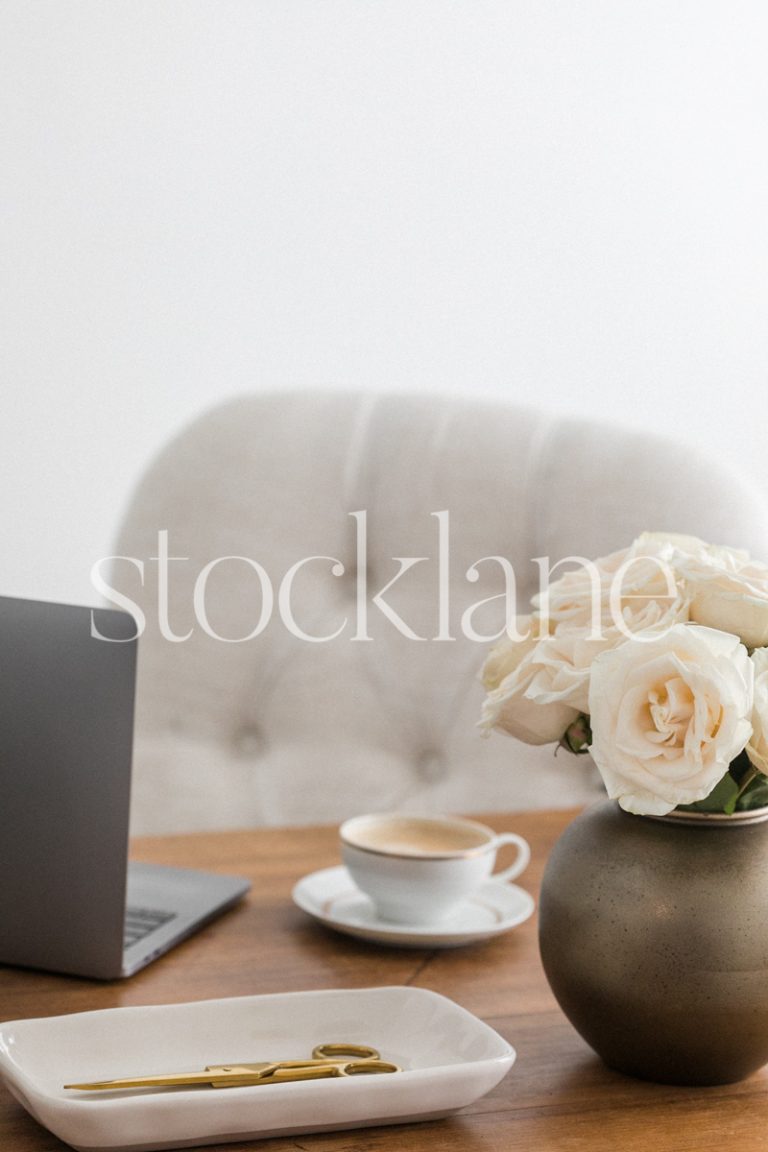 Vertical stock photo of a desk with a laptop computer, a vase with white roses, and a cup of coffee