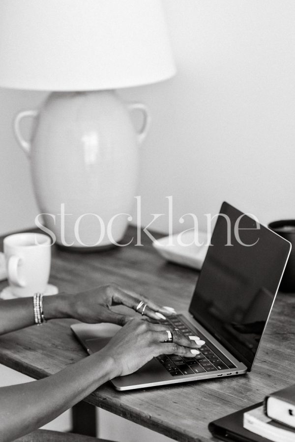 Vertical black and white stock photo of a woman sitting at her desk working on her laptop computer.