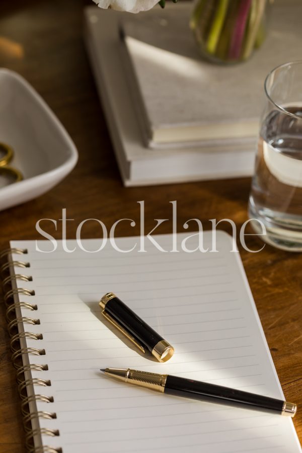 Vertical stock photo of a notebook and a pen on a wood desk, surrounded by other desktop items.