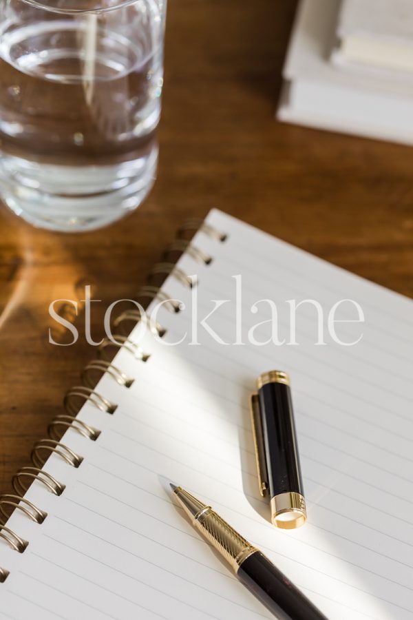 Vertical stock photo of a notebook and a pen on a wood desk, surrounded by other desktop items.