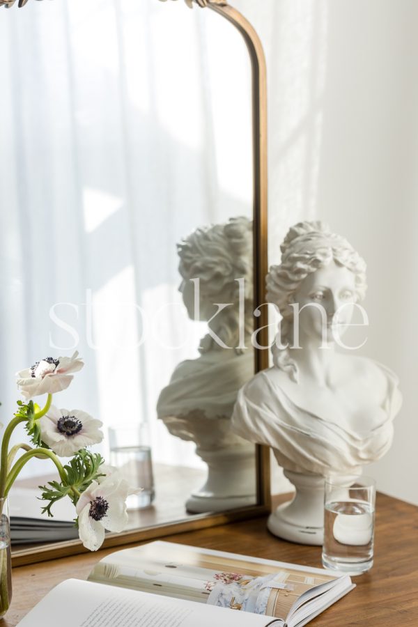 Vertical stock photo of a wood table with a mirror, sculpture, flowers and a magazine.